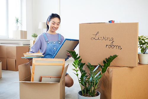 Full length portrait of young Asian woman packing decor items to cardboard boxes and smiling happily excited for moving to new house or dorm, copy space