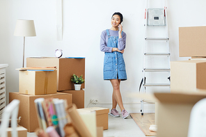 Full length portrait of young Asian woman speaking by phone and smiling happily while standing in empty white room with cardboard boxes, moving and relocation concept, copy space