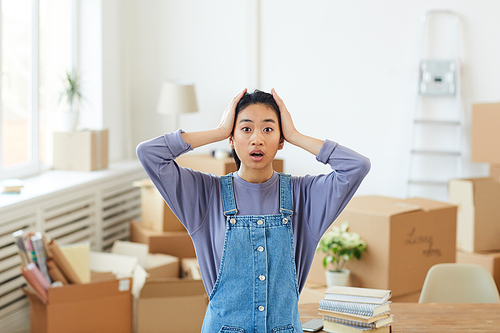 Waist up portrait of young Asian woman panicking while standing among cardboard boxes in empty room and  with big eyes, house moving or relocation concept, copy space