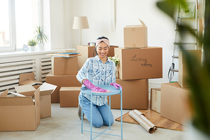 Full length portrait of cheerful Asian woman cleaning furniture while moving into new house with boxes in background, copy space