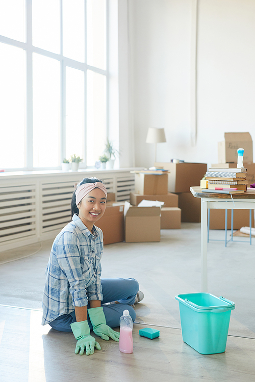 Vertical full length portrait of happy Asian woman sitting on floor in new house or apartment while cleaning after moving in, copy space