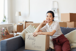 Portrait of young Asian woman smiling at camera while leaning on cardboard box with ready to move inscription, copy space