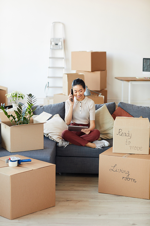 Vertical full length portrait of modern Asian woman using speaking by phone while unpacking boxes in new house or apartment, copy space