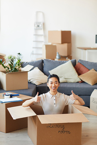 Vertical portrait of young Asian woman sitting in box and showing thumbs up while moving in to new house or apartment, copy space