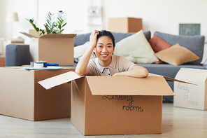 Portrait of young Asian woman sitting in box and smiling at camera while moving in to new house or apartment, copy space