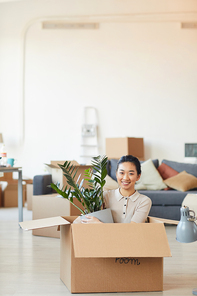 Vertical portrait of young Asian woman sitting in box and holding plant while moving in to new house or apartment, copy space