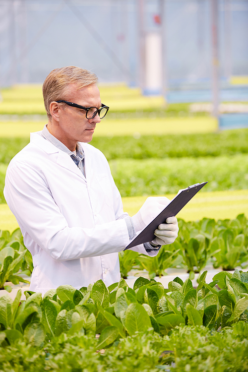 Vertical portrait of mature scientist holding clipboard while working on research at flower plantation in industrial greenhouse