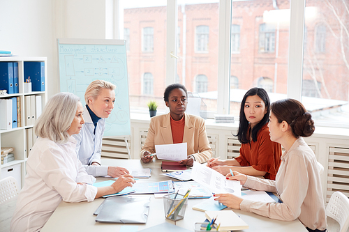 Multi-ethnic group of successful businesswomen discussing project while sitting at table against window during meeting in conference room, copy space