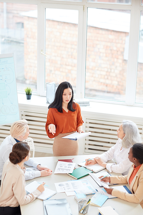Vertical portrait of successful Asian businesswoman presenting project plan to group of female colleagues while standing by table during meeting in conference room