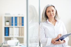 Waist up portrait of long-haired senior businesswoman  while holding clipboard and leaning against glass wall in office, left side copy space