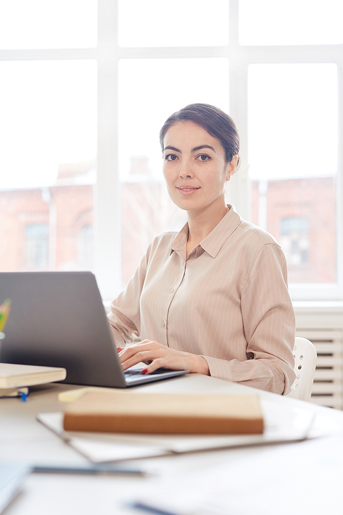 Vertical portrait of young elegant businesswoman  while using laptop sitting in white office against window, copy space above