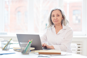Portrait of successful senior businesswoman  while using laptop sitting in white office against window, copy space