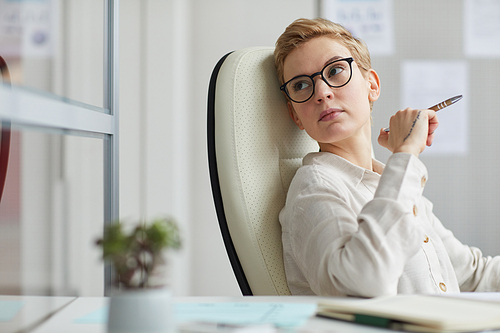 Portrait of modern short haired woman wearing glasses looking away pensively while sitting in big office chair at work, female boss concept, copy space
