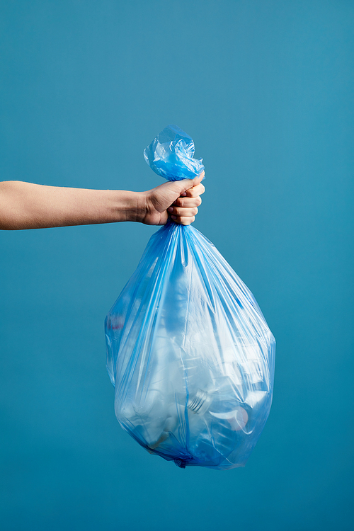 Vertical image of female hand holding trash bag with plastic against blue background, waste sorting and recycling concept, copy space