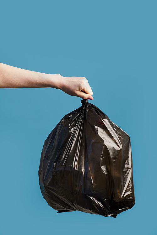Vertical image of female hand holding black trash bag against blue background, waste sorting and recycling concept, copy space