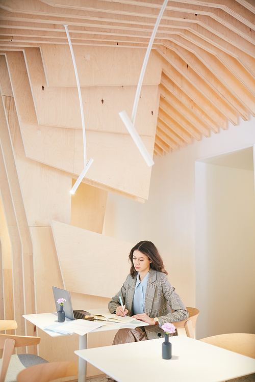 Vertical wide angle portrait of pretty young woman working or studying sitting at table in empty cafe hall with architectural design, copy space