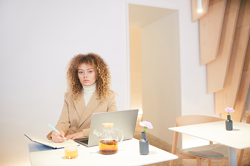 Warm-toned portrait of curly-haired young woman  while working or studying sitting at table in cafe hall, copy space