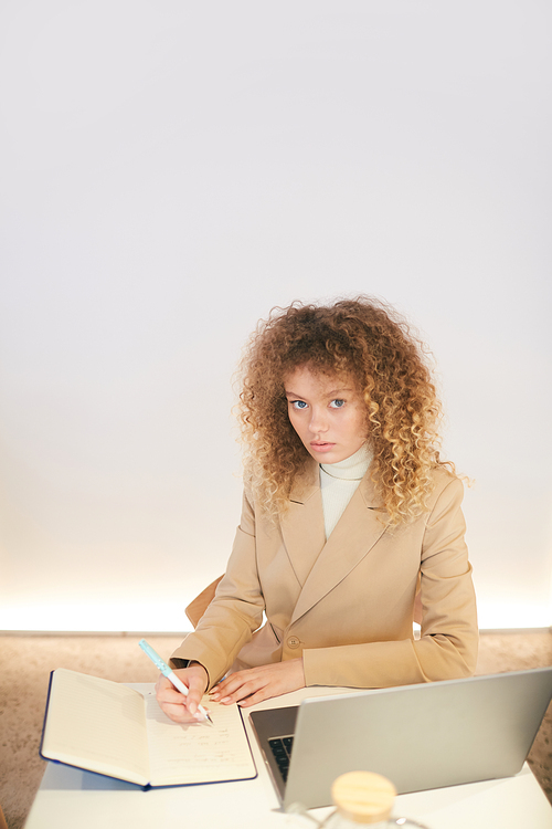 Vertical warm-toned portrait of curly-haired young woman  while working or studying sitting at table in cafe library hall, copy space