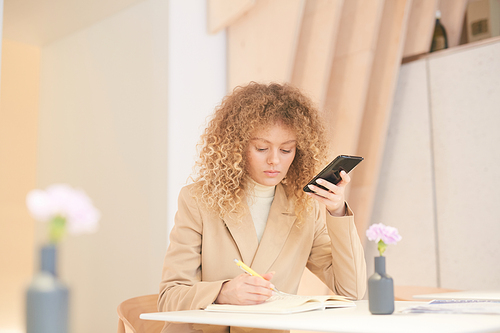 Warm-toned portrait of curly-haired young woman using smartphone while working or studying sitting at table in cafe, copy space