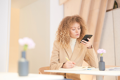 Warm-toned portrait of curly-haired young woman dictating message via smartphone while working or studying sitting at table in cafe, copy space