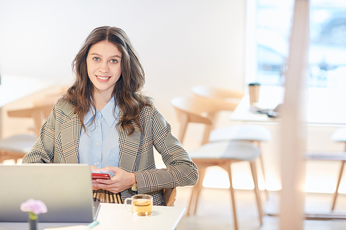 Candid portrait of pretty young woman  and holding smartphone while working or studying sitting at table in empty cafe, copy space