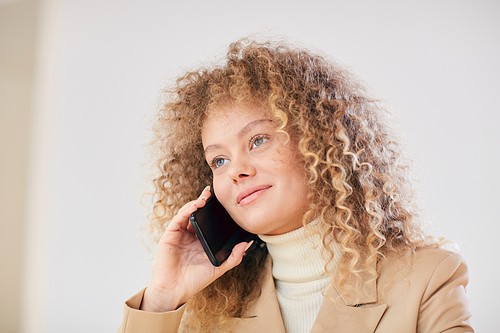 Head and shoulders portrait of curly haired young woman speaking by phone and smiling cheerfully while sitting against white wall, copy space