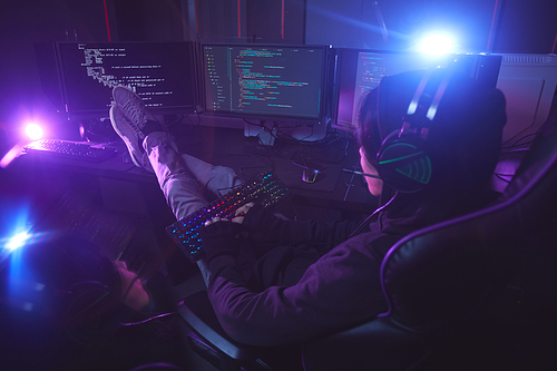 Back view at young man surrounded by multiple screens programming or hacking security in dark room, copy space