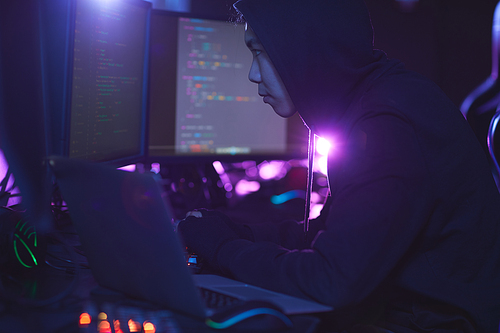 Side view at unrecognizable cyber security hacker wearing hood while working on programming in dark room, copy space