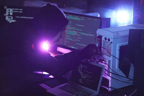 Side view at unrecognizable cyber security hacker wearing hood while working on computer system in dark room, copy space