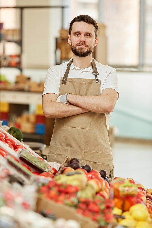 vertical waist up portrait of bearded man standing with arms crossed and  while selling fresh fruits and s at farmers market