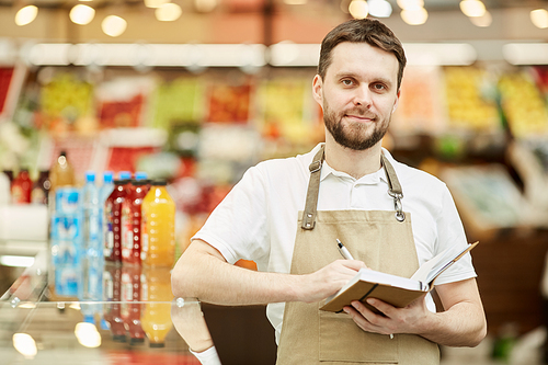 Waist up portrait of bearded man wearing apron and holding notebook while standing in supermarket, copy space