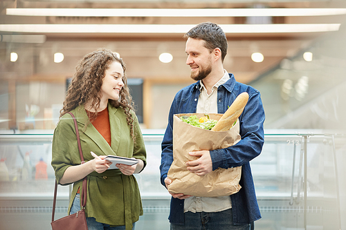 Waist up portrait of cheerful couple holding paper bag with groceries while enjoying shopping in supermarket, copy space