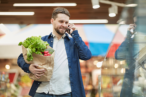 Waist up portrait of smiling bearded man calling by smartphone while grocery shopping in supermarket, copy space