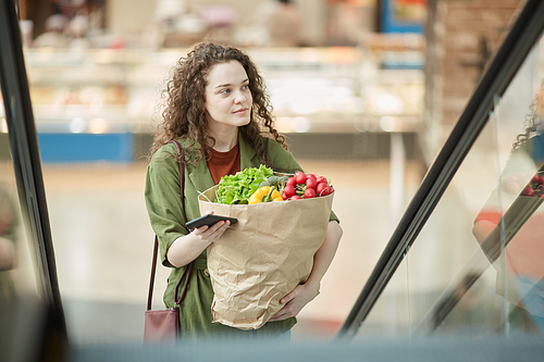 High angle portrait of modern young woman holding bag with groceries while going up escalator in mall, copy space