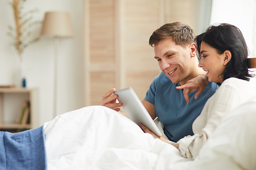 Side view portrait of smiling adult couple using digital tablet in bed in home interior while choosing online services, copy space