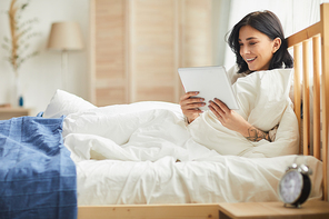 Portrait of smiling adult woman using digital tablet and browsing internet while lying in bed in morning, copy space