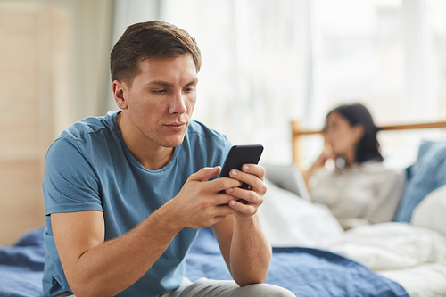 Portrait of adult man using smartphone while sitting on bed at home with wife in background, family fight, communication problem concept