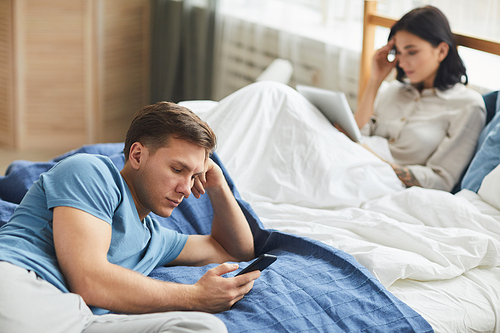 Portrait of modern young couple using mobile devices while sitting on bed, communication problem concept, copy space