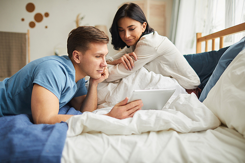 Portrait of modern young couple using digital tablet while sitting on bed at home and ordering food online or watching movie, copy space