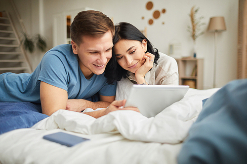 Portrait of young happy couple using digital tablet while lying on bed at home and ordering food online or watching movie, copy space