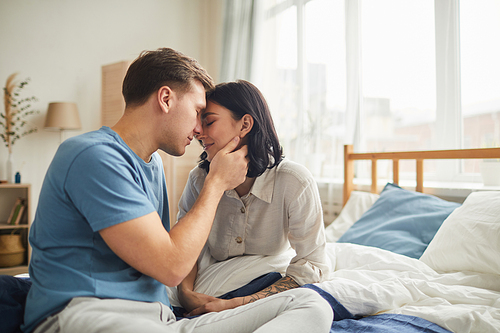 Side view portrait of loving young couple kissing tenderly while sitting on bed in morning, copy space