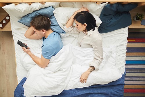 Top view portrait of modern young couple sleeping together with man using smartphone next to wife, copy space