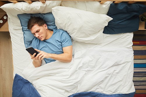 Above view portrait of modern young man using smartphone while lying on bed in blue and white interior, copy space