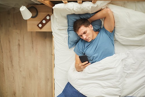 Top view portrait of modern young man using smartphone while lying on bed in blue and white interior, copy space