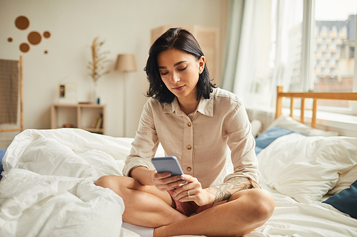 Warm-toned full length portrait of modern young woman using smartphone while sitting on bed in morning, copy space