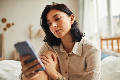 Warm-toned portrait of modern young woman using smartphone while sitting on bed in morning, waiting for text message, copy space