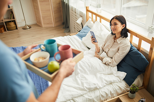 High angle view at unrecognizable caring man bringing breakfast in bed for young woman holding smartphone, warm-toned family scene, copy space