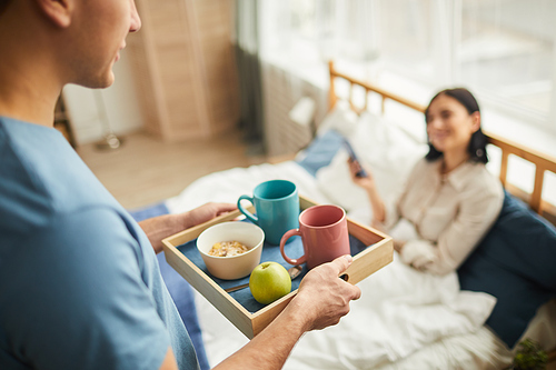 High angle close up of unrecognizable caring man bringing breakfast in bed for young woman holding smartphone, warm-toned family scene, copy space