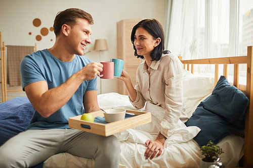 Side view portrait of happy young couple enjoying breakfast in bed and clinking coffee mugs while looking at each other with love, copy space