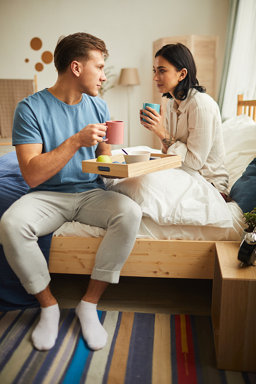 Full length portrait of happy young couple enjoying breakfast in bed and clinking coffee mugs while looking at each other with love
